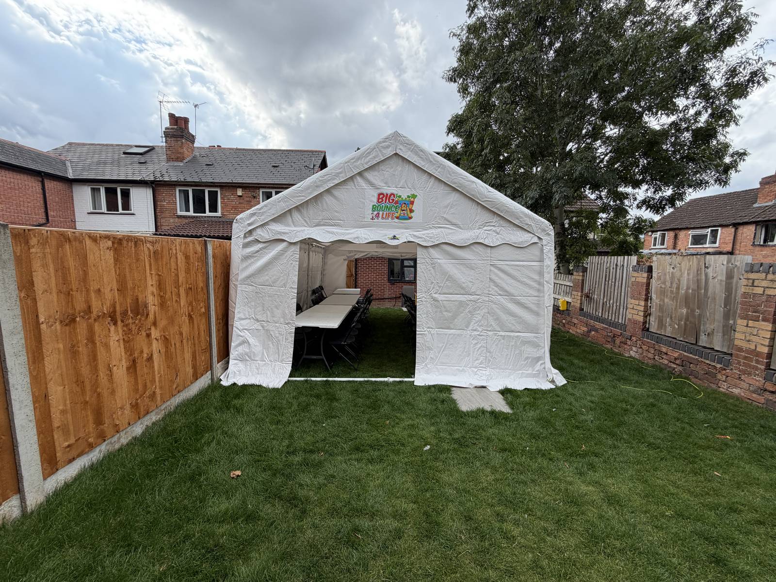 Small white marquee on a neat lawn against a fence, with a brick house behind