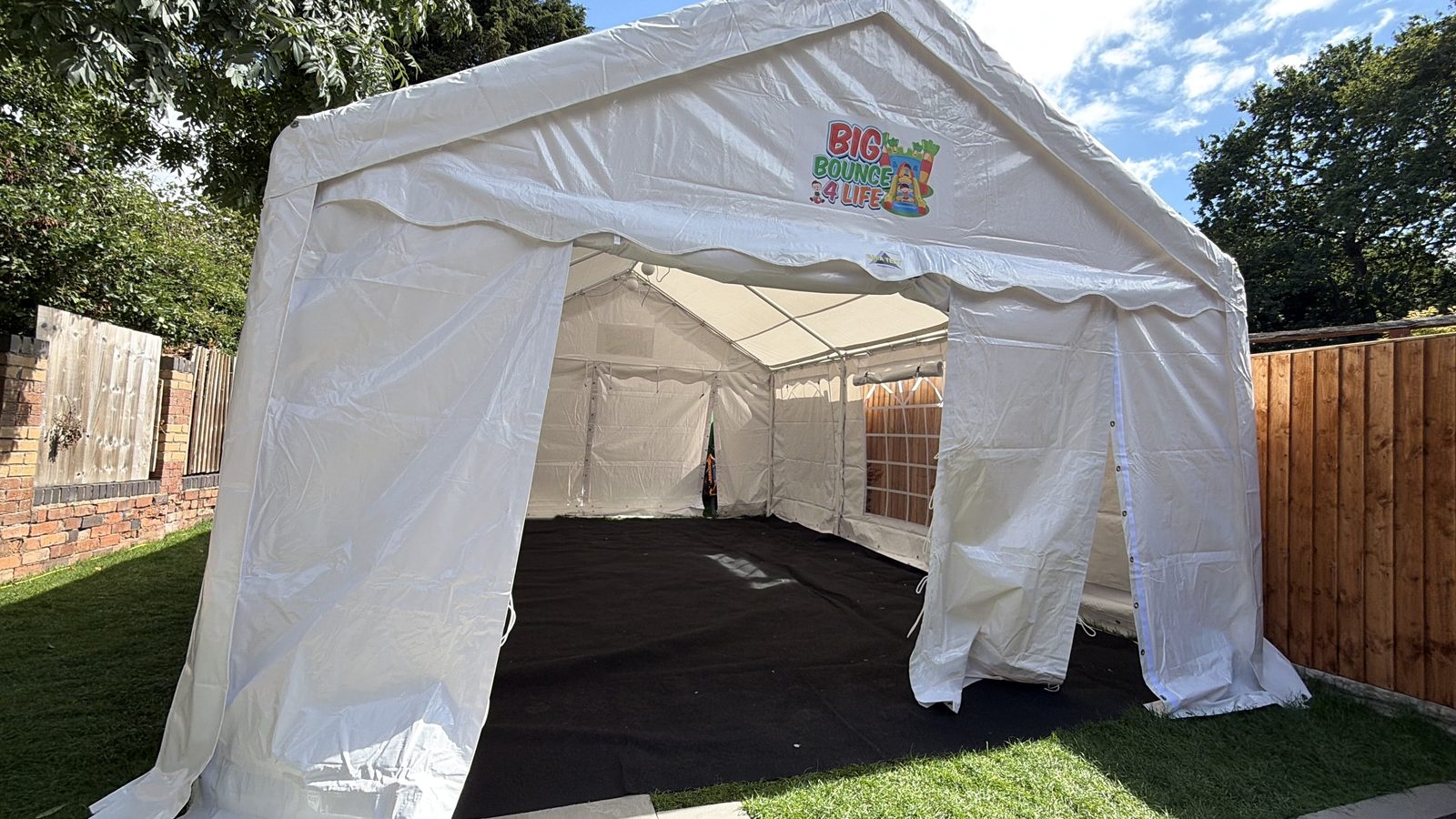 Open-fronted marquee with side panels up and black flooring on a garden lawn